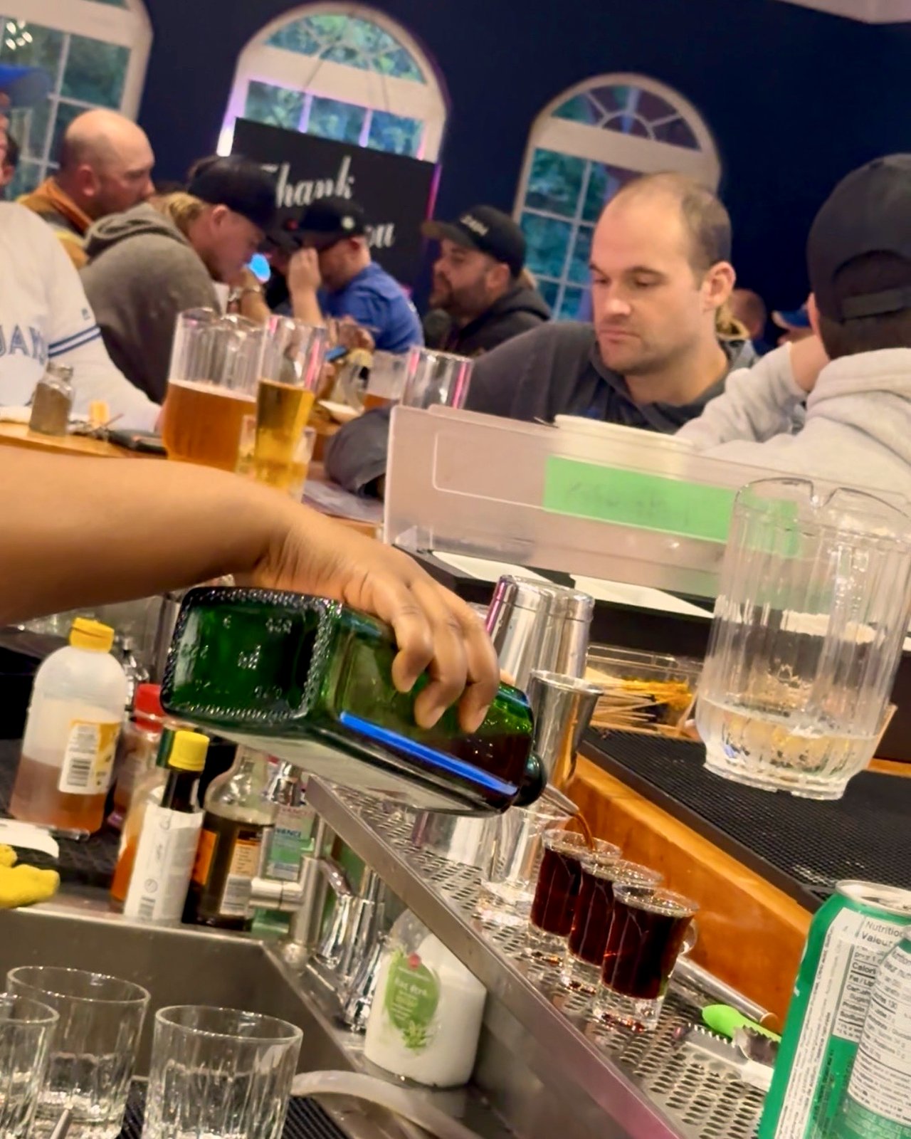 Bartender pouring shots behind the bar with a packed crowd of Toronto Blue Jays fans watching the game at OV Burger Bar