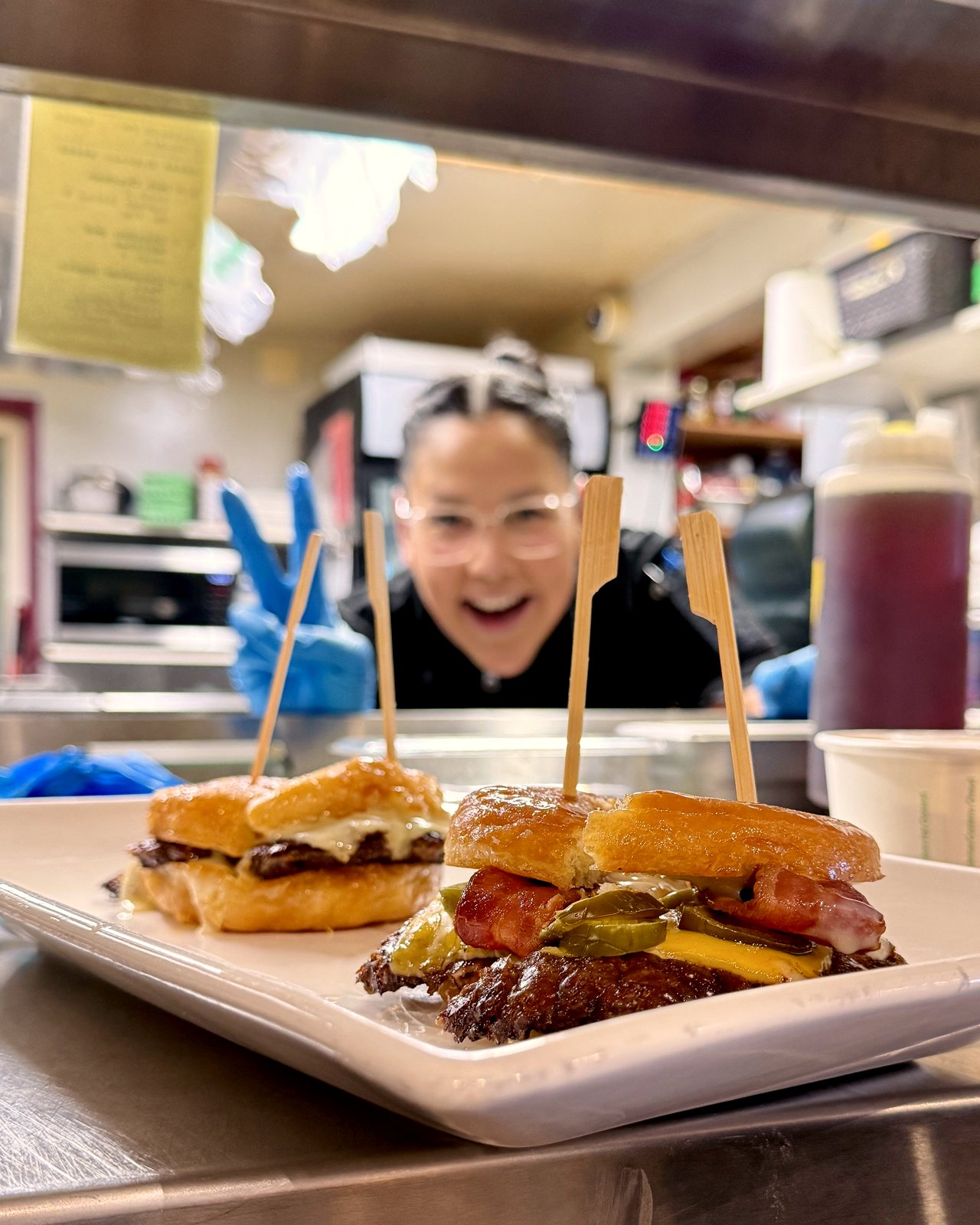 Bonnie, OV's morning kitchen manager, throwing a peace sign behind two smash burgers at the pass at OV Burger Bar in Prince Rupert, BC