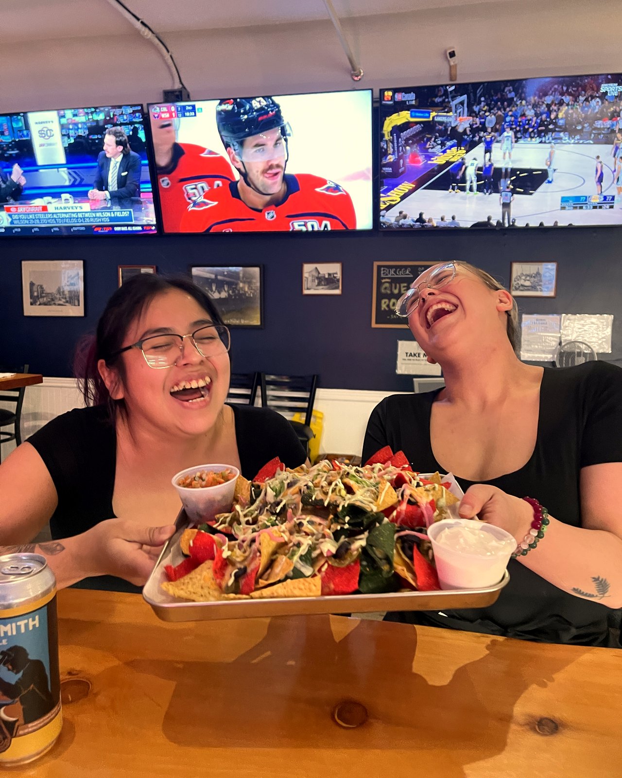 Two OV servers laughing while presenting a tray of OV Game Day Nachos under the sports TVs at OV Burger Bar in Prince Rupert, BC