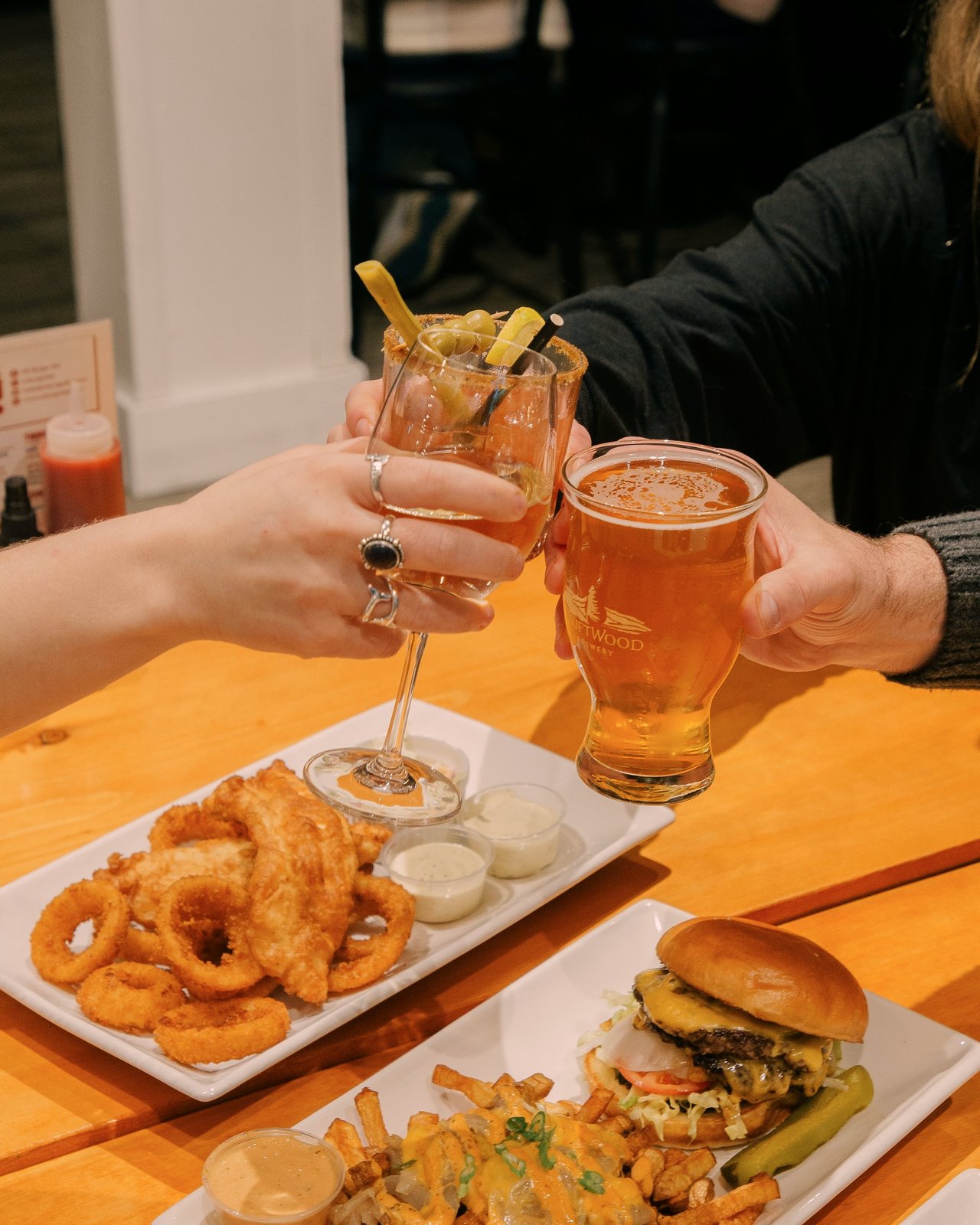 Two guests toasting a cocktail and a craft beer over a table of food at OV Burger Bar in Prince Rupert, BC