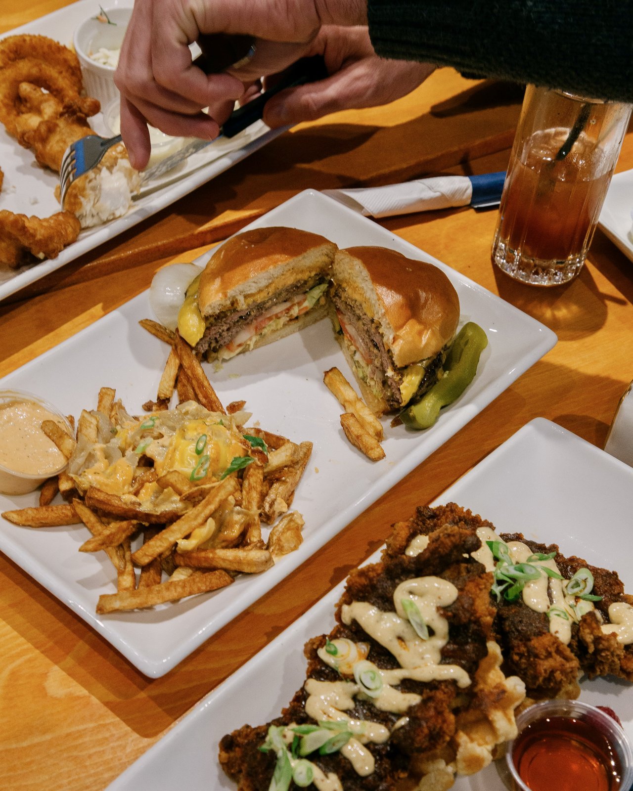 A smashed cheeseburger cut in half showing the patty and melted cheese, animal fries, and chicken and waffles on the table at OV Burger Bar in Prince Rupert, BC. Someone takes a photo of their meal in the background.