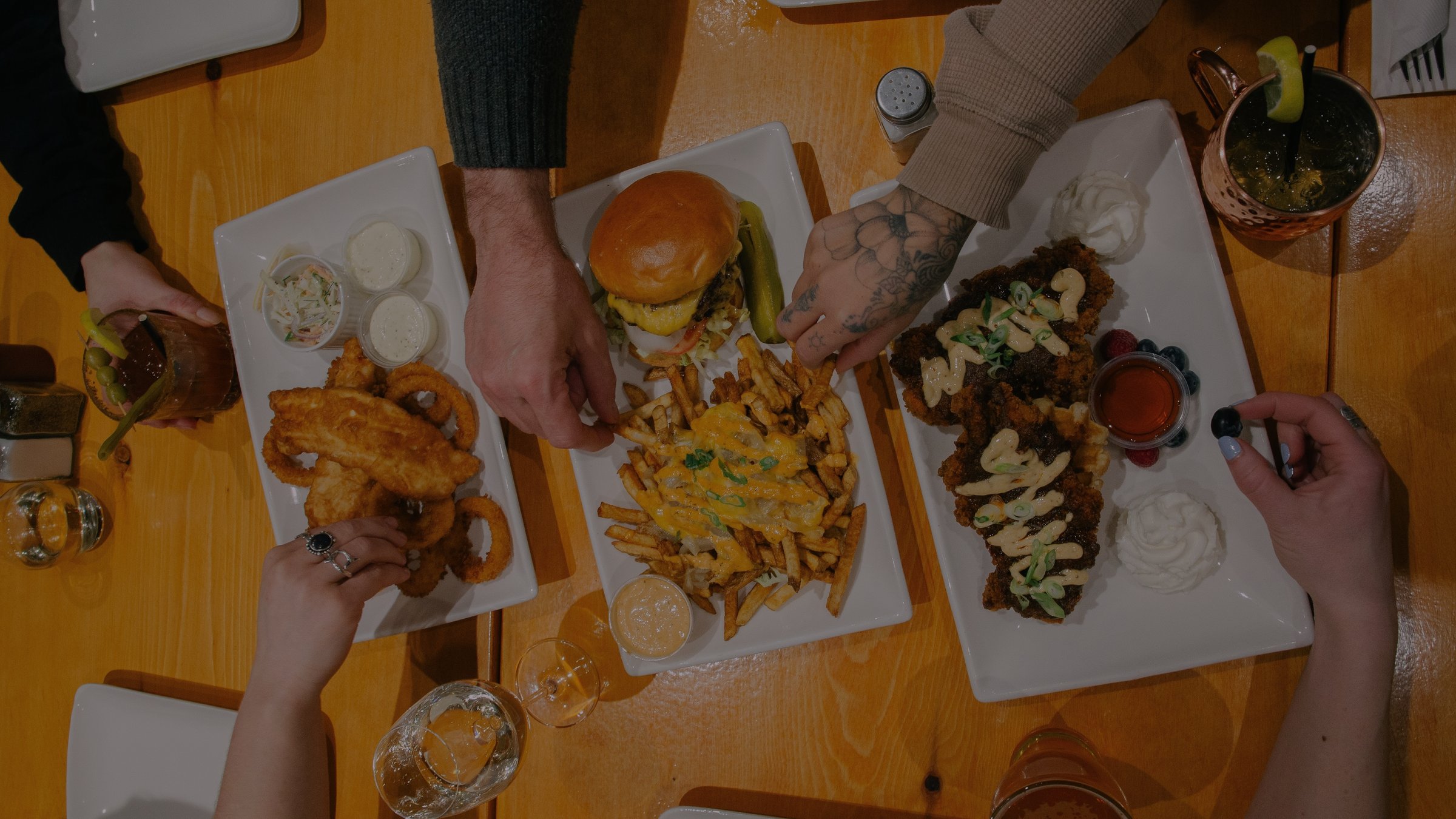 Three plates of shareable food being shared at a wooden table at OV Burger Bar in Prince Rupert, BC. A double cheeseburger and animal fries in the centre, fish and chips with onion rings on the left, and chicken and waffles with maple syrup on the right. Hands reaching in, beer and a Moscow mule visible.